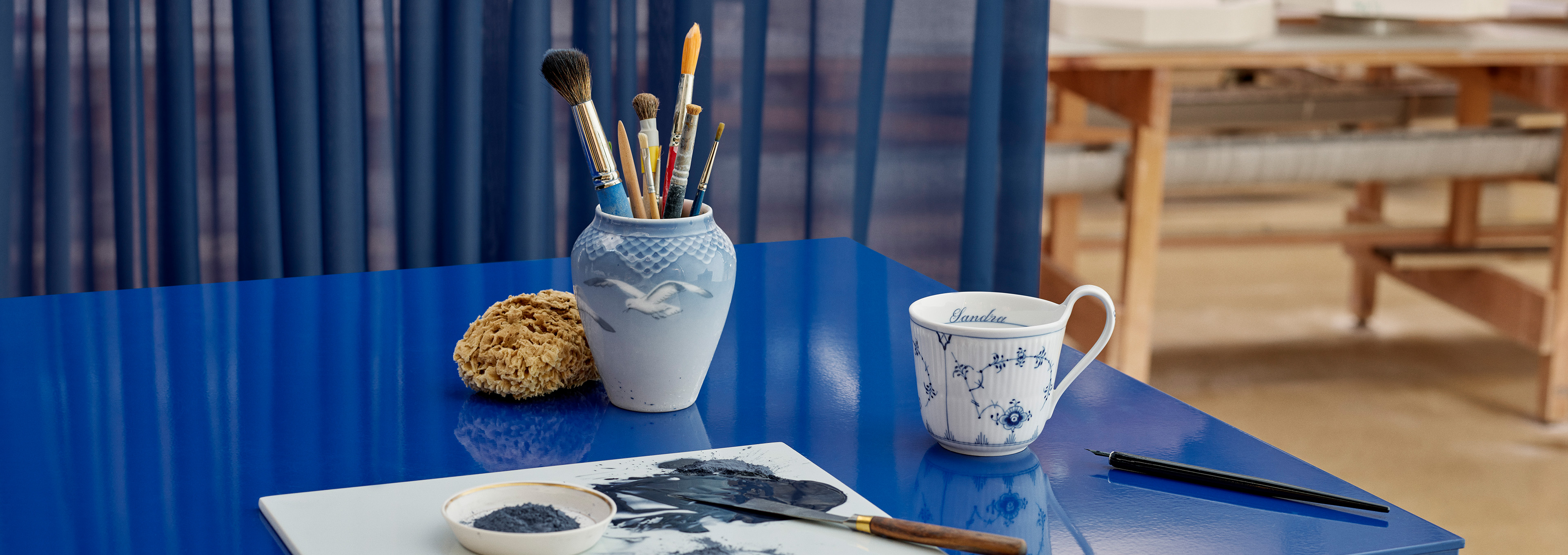 A Blue Fluted Plain High Handle Cup 33 cl with a freshly painted name on the inside standing on a blue desk next to brushes and paint